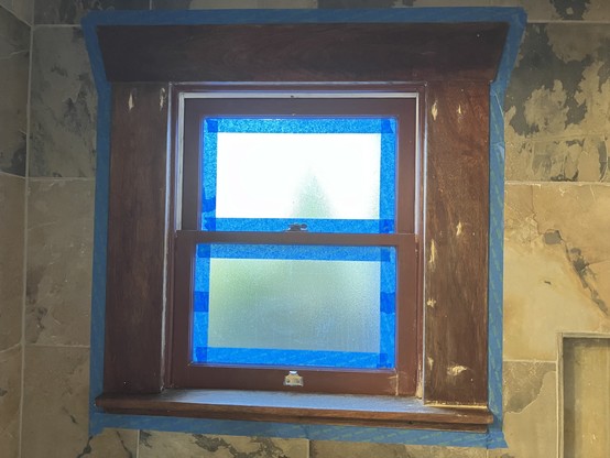 A darkly brownish colored window with frosted panes, surrounded by slate tile and painter’s tape. There is wood putty in spots.